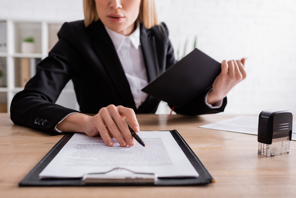 Kobieta w biznesowym stroju siedzi przy biurku i z długopisem w dłoni przegląda dokumenty