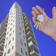 Nowy, wysoki apartamentowiec. Na pierwszym planie dłoń trzymająca kluczyki z brelokiem w kształcie domu