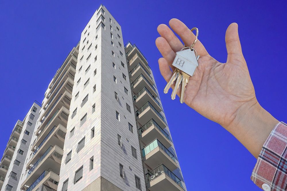 Nowy, wysoki apartamentowiec. Na pierwszym planie dłoń trzymająca kluczyki z brelokiem w kształcie domu