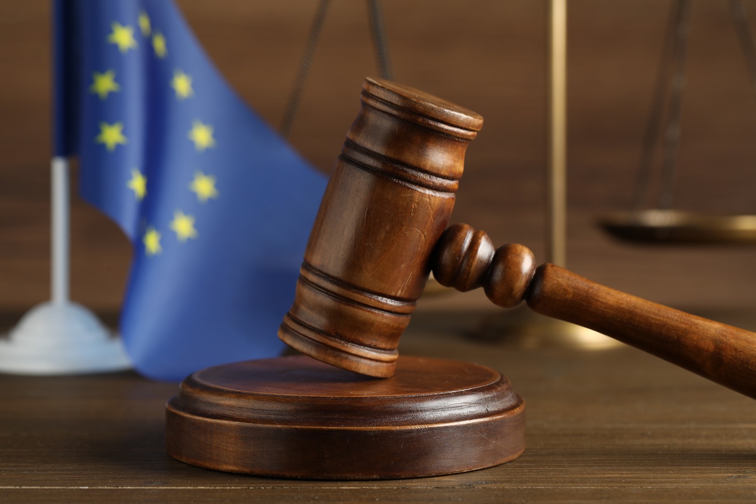 Drewniany blat, na nim sędziowski młotem z podstawką, w tle flaga Unii Europejskiej i waga, symbolizująca sprawiedliwość