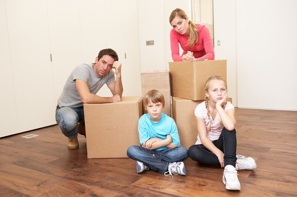 Zmartwiona rodzina (rodzice i dwoje dzieci) siedzi w pustym mieszkaniu, z ustawionymi kartonowymi pudełkami